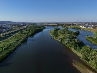 Fototapeta premium Aerial view of calm river winding through green landscape. Blue sky stretches above serene waterway and trees. Urban skyline blends with natural scenery in distance. Ideal for environmental, travel