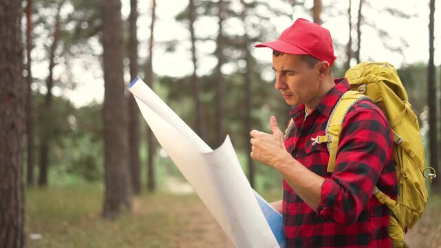 Hiker reading map in pine forest. Man with backpack navigates trail among tall trees. Traveler studies route on paper map. Hiker searches direction in forest. Backpacker plans hiking path.