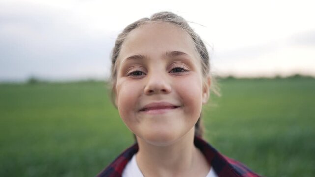 Cute girl looking at camera. Child stands in green field. Outdoor portrait shows innocent face. Young girl gives sweet smile. Happy child enjoys beautiful nature. Smiling face shows real emotion.