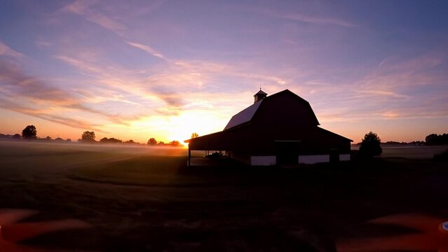 Rural barn silhouette at sunrise over misty farmland, warm golden sky and soft fog across fields, peaceful countryside landscape for agriculture and nature themes