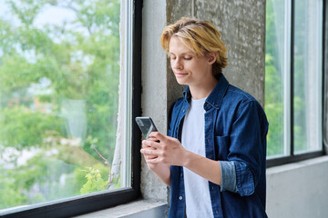 Fototapeta premium Smiling teenage boy using a smartphone by window