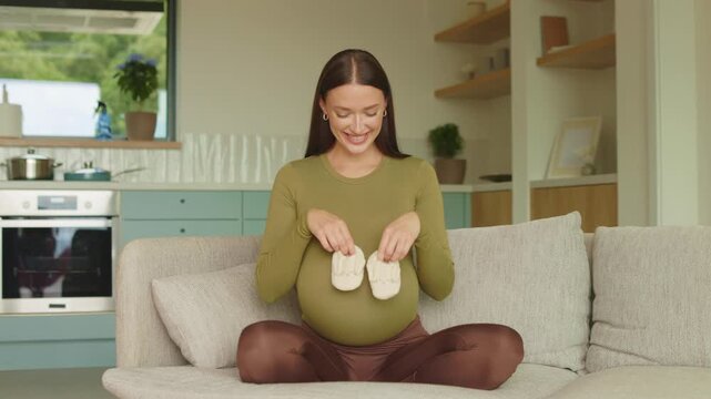Pregnant woman smiling while playing with tiny baby shoes at home