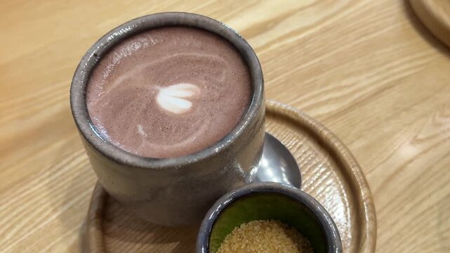 Hot cocoa with heart foam art on wooden coaster beside cane sugar and teaspoon