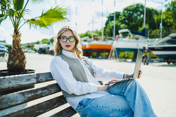Fototapeta premium Woman works on laptop while sitting on bench near boats during sunny day in marina