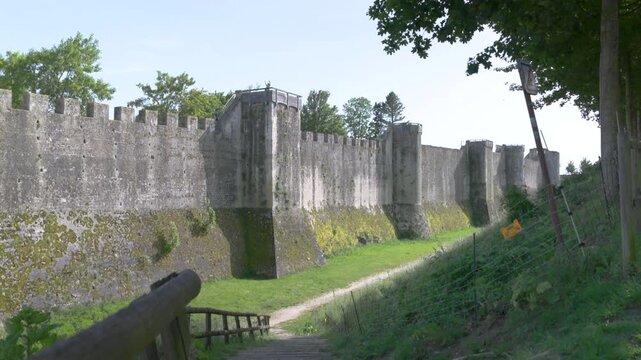 Imposing ancient stone fortress wall featuring fortification towers and a grassy defensive moat on a sunny day.