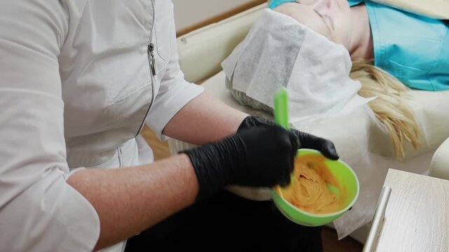 A female doctor, a professional cosmetologist, mixes a mask for application to a patient's face