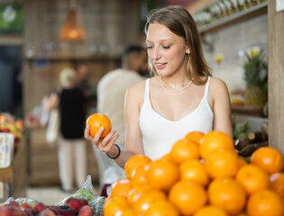 Fototapeta premium Young blonde woman chooses ripe oranges in the grocery section of a supermarket. Visitors to the fruit section buy citrus fruits and fruits