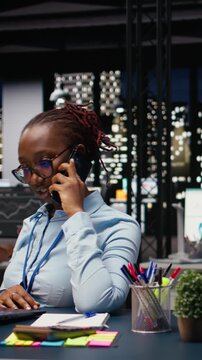 Vertical Video African american woman answering phone call for a briefing discussion, using laptop and requesting assistance. Manager does research on profitability, telephone call in office.