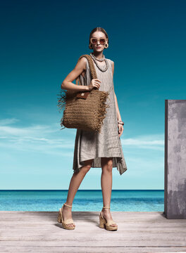 Exquisite high fashion portrait of stunning Caucasian model wearing beige linen dress woven wedges and holding large fringed straw shoulder bag on wooden pier at exotic tropical beach resort