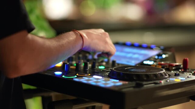 Hand of a disc jockey turning a knob on a glowing dj controller with backlit performance pads jog wheel and screen during a live music set