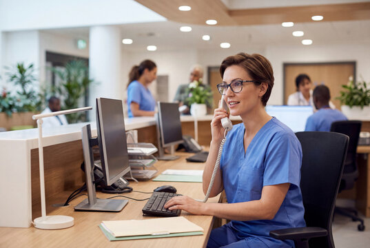 Female hospital receptionist in blue scrubs and glasses talking on telephone at front desk