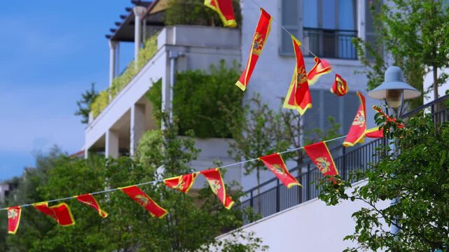 Montenegro national flag strung as festive bunting fluttering outdoors near a white building and green tree, patriotic street decoration, national holiday mood, summer celebration, Statehood Day