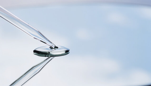 Glass dropper pipette dispensing clear fluid drop on reflective mirror surface with sky reflection