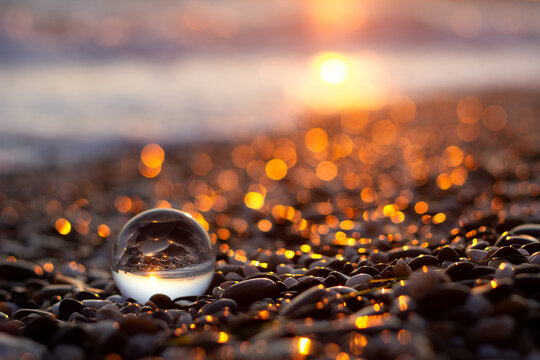 Artistic closeup of a crystal lensball on a pebble beach at golden sunset, reflecting upside down sea horizon with bokeh background.