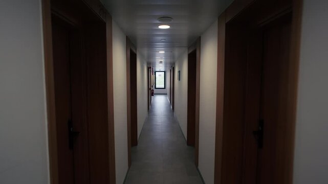 Point of view moving along a long narrow corridor lined with wooden doors and white walls leading toward a bright window at the end