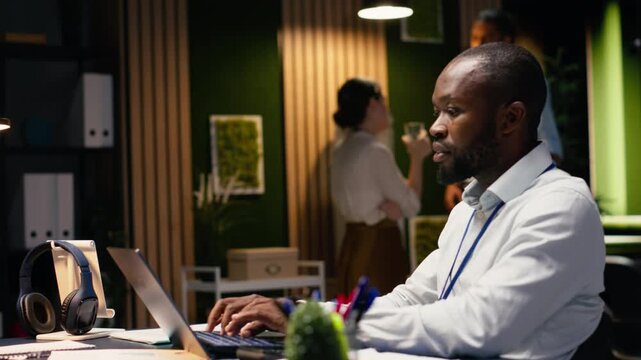 Black man accountant in office using laptop and writing down information, working late to finish tasks before deadline. Transcribing from screen and taking notes, documenting research.