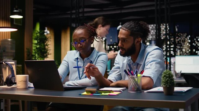 Smiling business associates in office evaluating staff performance with laptop, pleased after completing task at night. Team focused on documenting business intel, reviewing positive metrics.