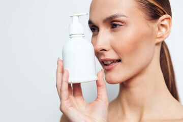 Young woman with clear skin holds white pump bottle close to face, looks at product with slight...