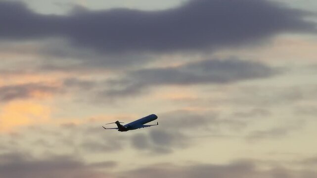 Silhouette of a small regional jet climbing through pastel clouds at sunset with warm golden and grey tones filling the evening sky