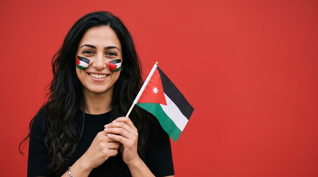 Happy young woman with Jordanian flag painted on her cheeks holding a small national flag of Jordan against a vibrant red background