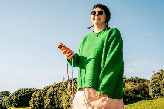 Woman Stands in Park Wearing Green Sweater and Holding Phone