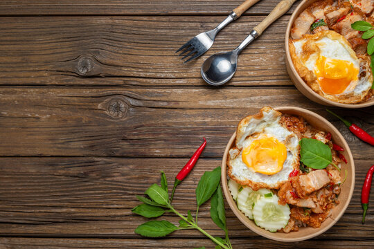 Thai street food, Pad Kra Pao Moo Krob, spicy crispy pork with basil and fried egg served in takeaway bowls on wooden table.
