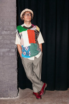 Man stands in a studio wearing a colorful sweater and gray pants