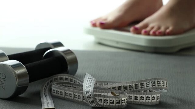 Woman standing on scales indoors, focus on measuring tape, dumbbells and fitness mat