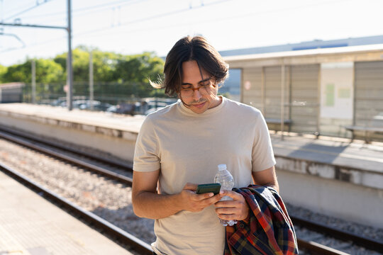Young man using smartphone and holding water bottle at train station