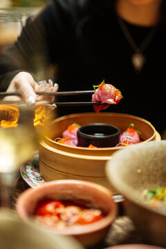 Person holding pink dumpling with chopsticks over bamboo steamer