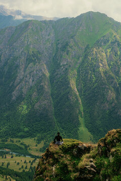 Person sits on cliff edge overlooking green mountains and valley