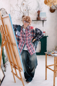 Artist stands by easel in a bright studio with supplies