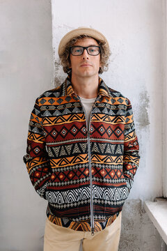 Man poses in patterned jacket with glasses and hat by the wall