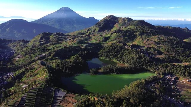Aerial view of the vibrant green Telaga Warna lake surrounded by terraced hills and the majestic Mount Sindoro peak under a clear blue sky in Indonesia.