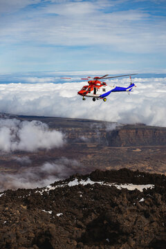 Red rescue helicopter flying above mountains and clouds.