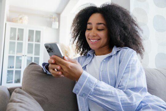 Young smiling black woman watching smartphone on sofa at home