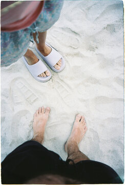 Two pairs of feet on sand at the beach with one wearing slippers