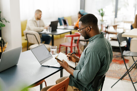 Young Man Writing Notes in Cafe While Using Laptop and Notebook