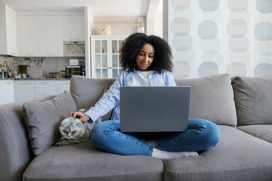 Black woman caressing cat while watching laptop on couch at home