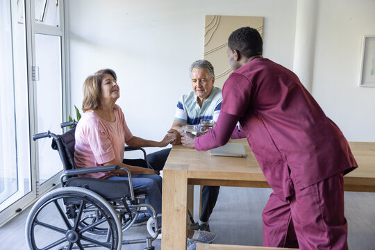 Diverse senior woman in wheelchair holding hand, aide in maroon scrubs placing pitcher on table