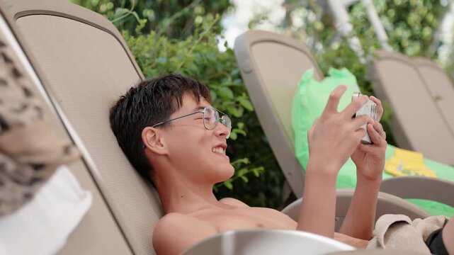 Teenage traveler relaxing on sun lounger and using smartphone near resort pool during sunny family vacation. Digital lifestyle and summer leisure concept
