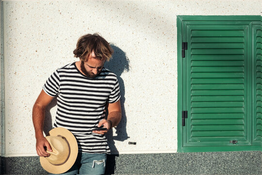 Young man holding hat and smartphone texting by green shutter