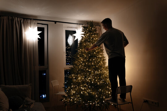 Man standing on chair decorating christmas tree with lights at home