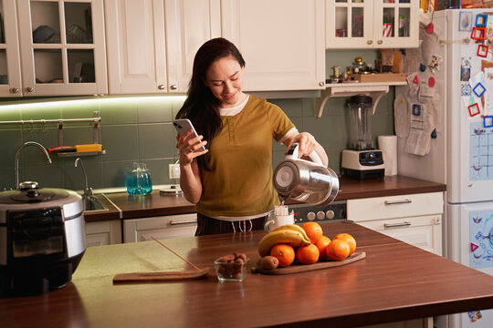 Woman makes tea in kitchen while using smartphone