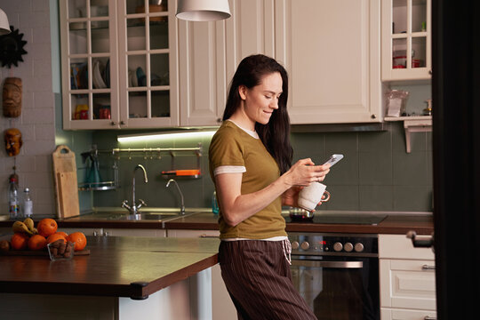 Woman enjoying coffee and using smartphone in kitchen