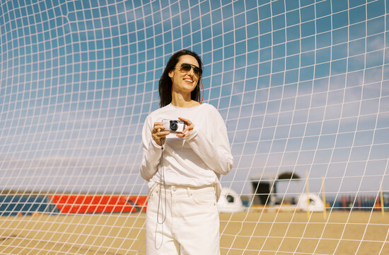 Woman Holds Camera Near Net Structure on Beach in Bright Daylight