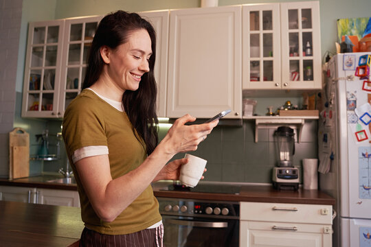 Woman enjoying coffee and using smartphone in kitchen