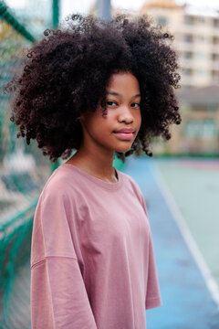Teen with curly hair poses by tennis court fence.