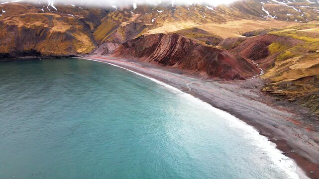 Aerial view of Brunavik bay with its reddish cliffs and dark sand beach meeting turquoise ocean waves under a cloudy sky in Bakkagerdi, Mulathing, Iceland.