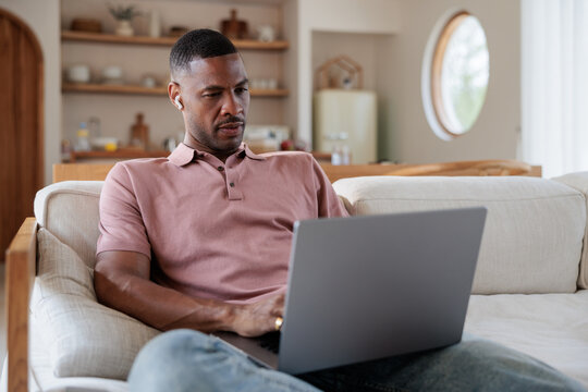 Man on couch works on laptop with earbuds in.
